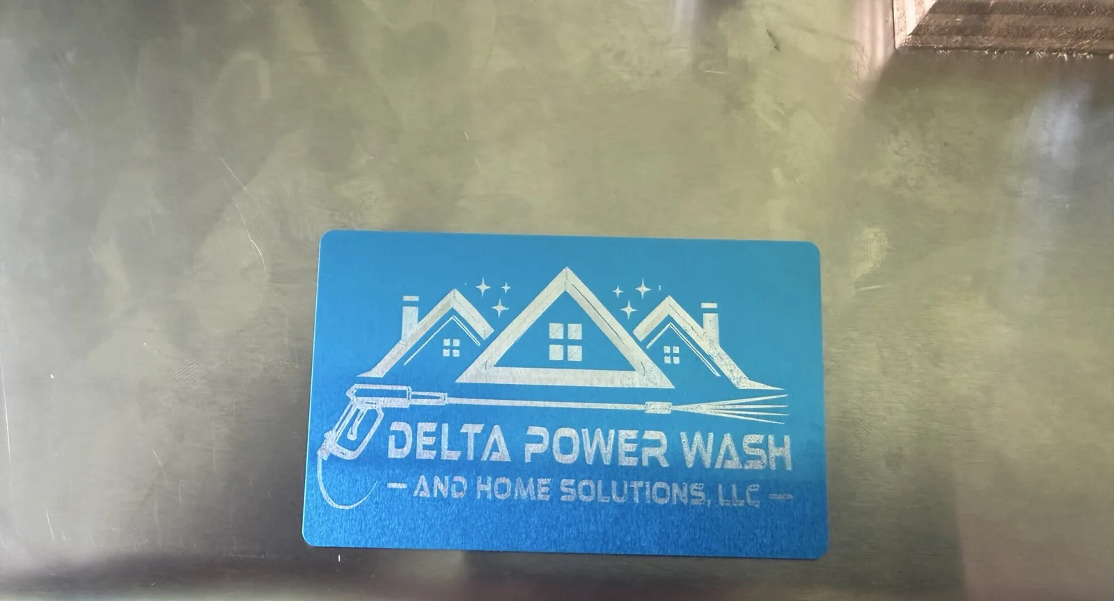 Blue brushed metal business card engraved for Delta Power Wash and Home Solutions LLC with house and pressure washer icon