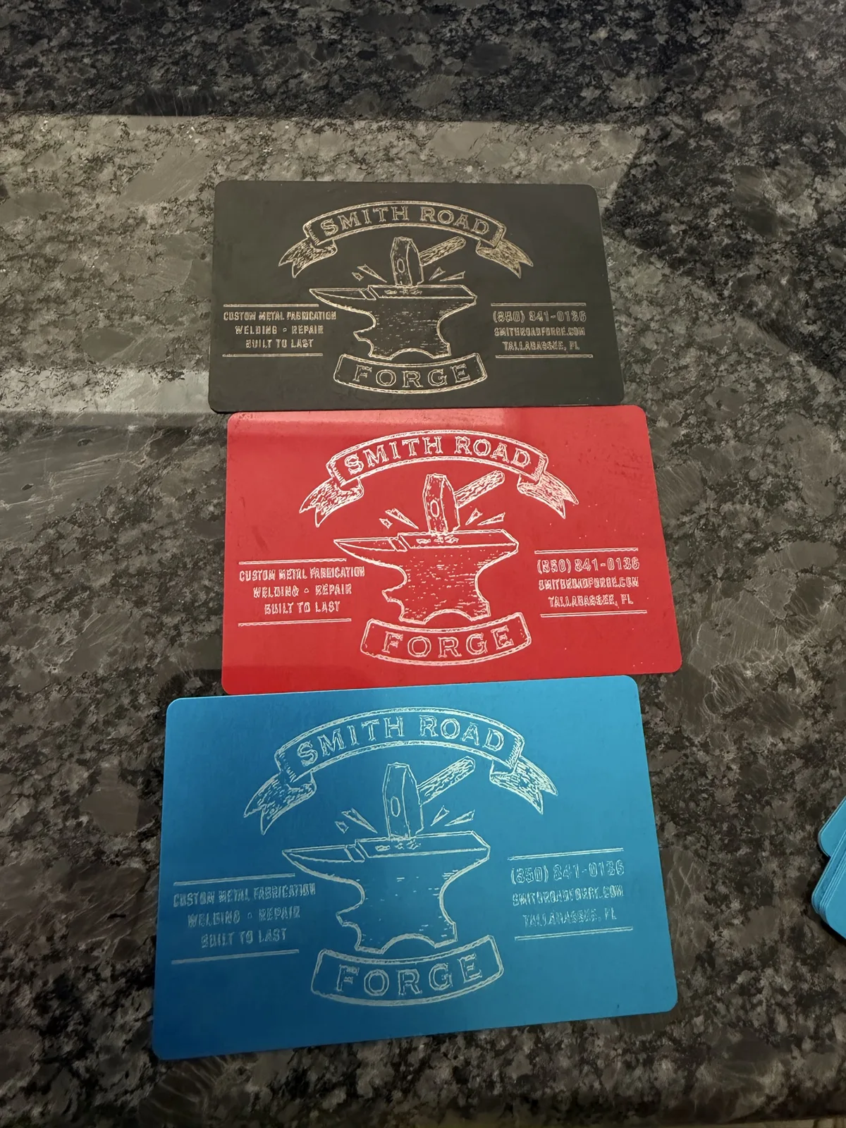 Three engraved metal business cards for Smith Road Forge in black, red, and blue finishes with anvil and hammer logo